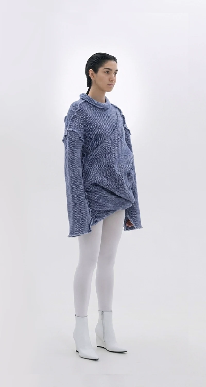 Model wearing the finished sculptural blue-gray sweater with white trousers and boots